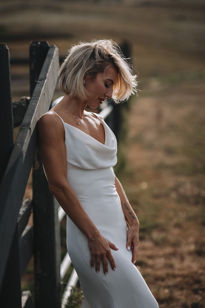 Elegant white slip wedding dress catches golden sunlight as a bride leans against a rustic wooden fence in a countryside setting.