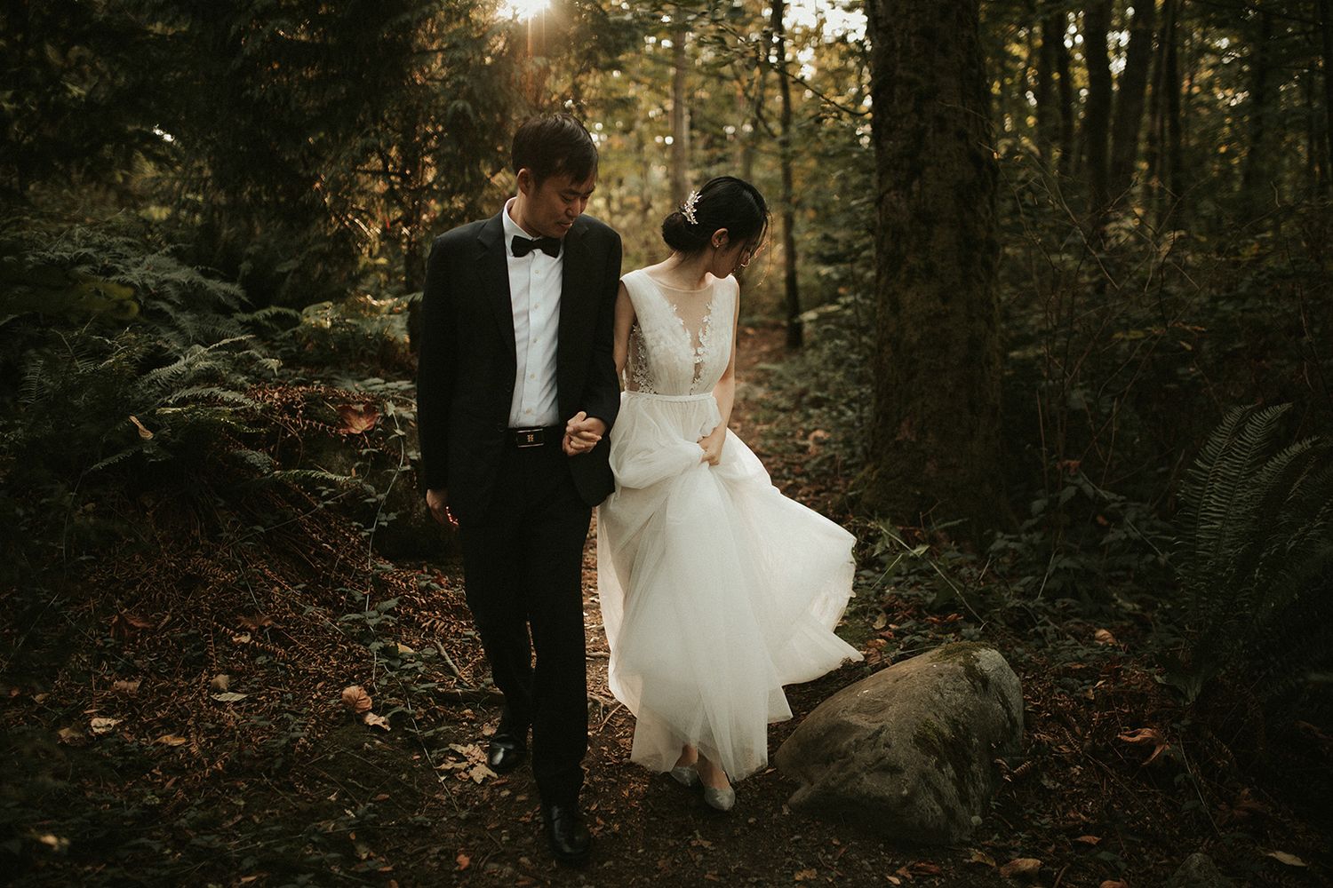 A couple in wedding attire walks through a sunlit forest path during golden hour. The bride looks back as though she dropped something.
