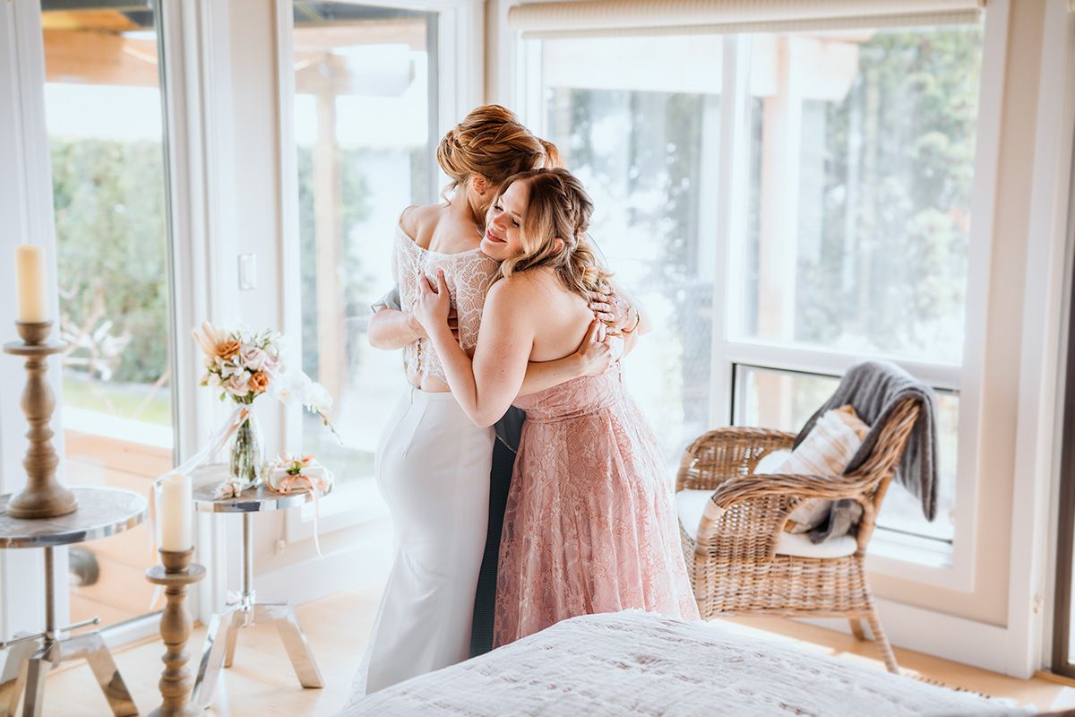 A bride and her sister embrace in a rustic interior with white curtains and vintage decor elements arranged by large windows.