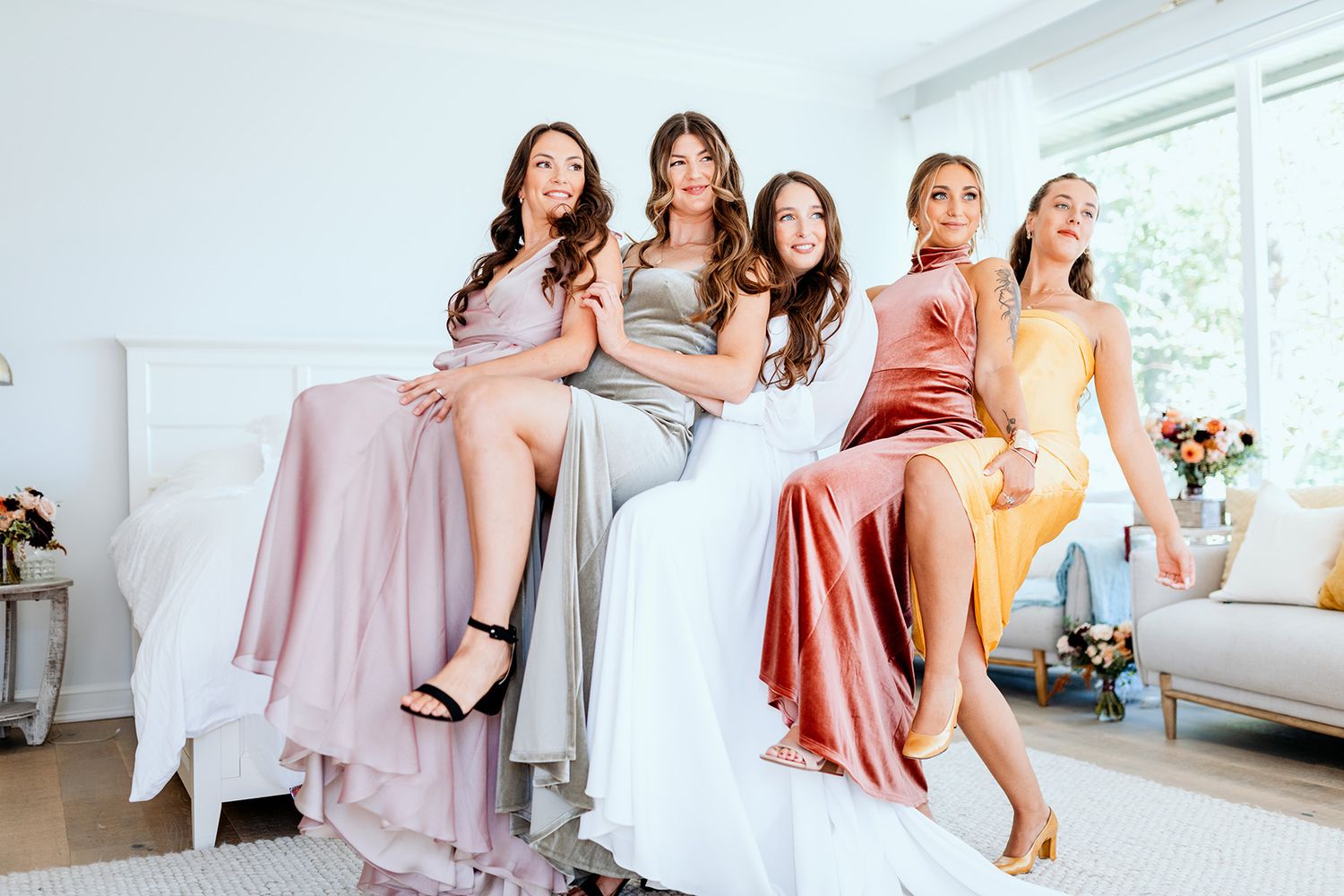 Bridesmaids in pink and coral dresses laugh and dance together in a bright bedroom with large windows.