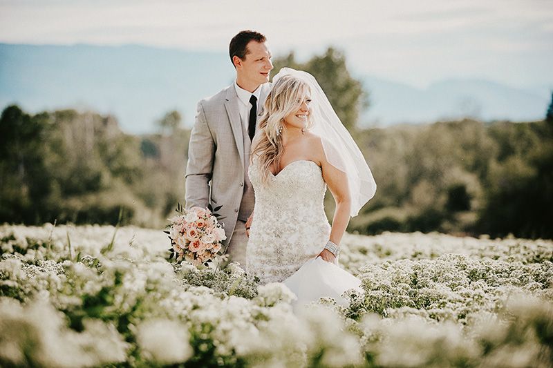 Couple stands in a dreamy meadow of white wildflowers on their wedding day as sunlight filters through the trees.