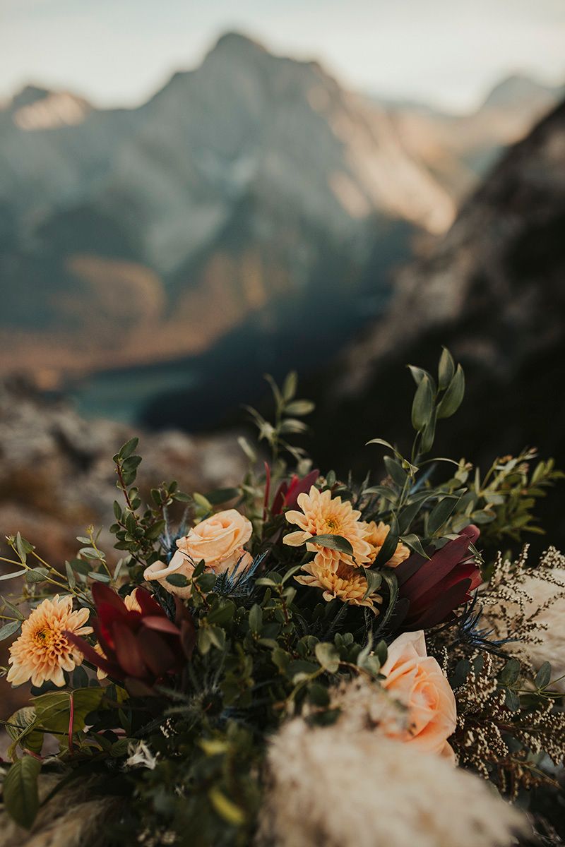 Peach and burgundy floral arrangement with greenery against a blurred mountain landscape backdrop at sunset.
