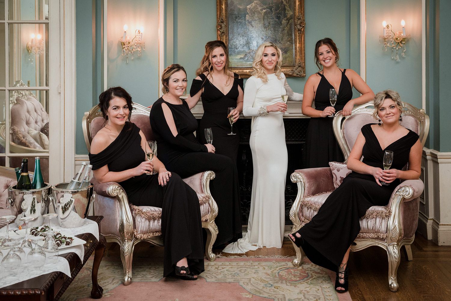 Wedding party in black dresses poses together in an elegant room with mint green walls and crystal chandeliers.