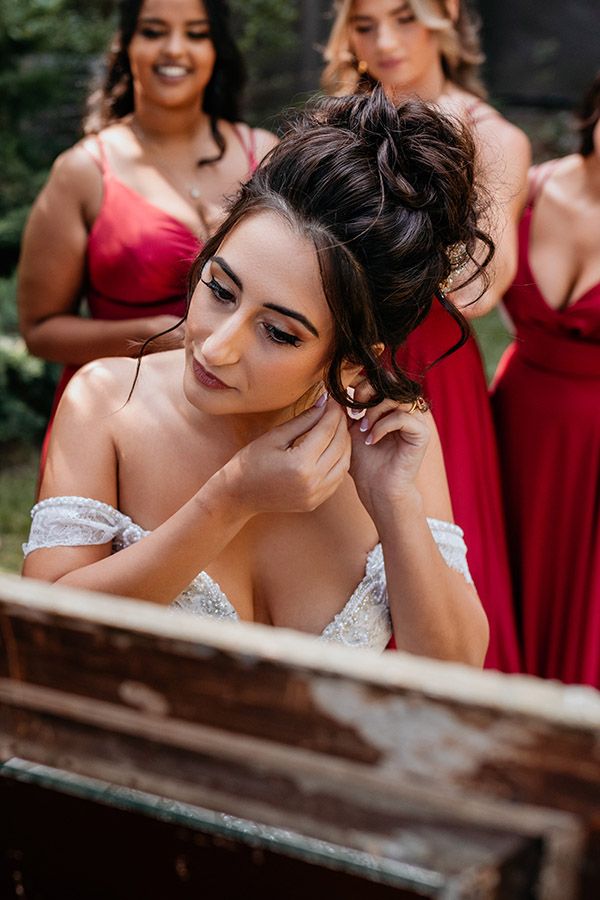 A bride looks into a mirror as she puts in her earrings while her bridesmaids in red dresses surround her..