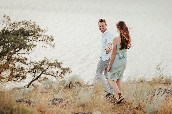 Couple holding hands smiling outdoors near a lake with tall grass and a tree in the background.