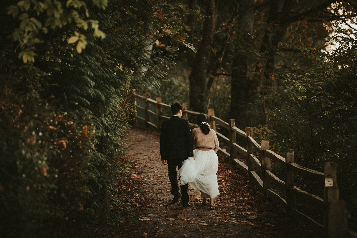 A couple in wedding attire walks along a rustic wooded path with a wooden fence on an autumn day.
