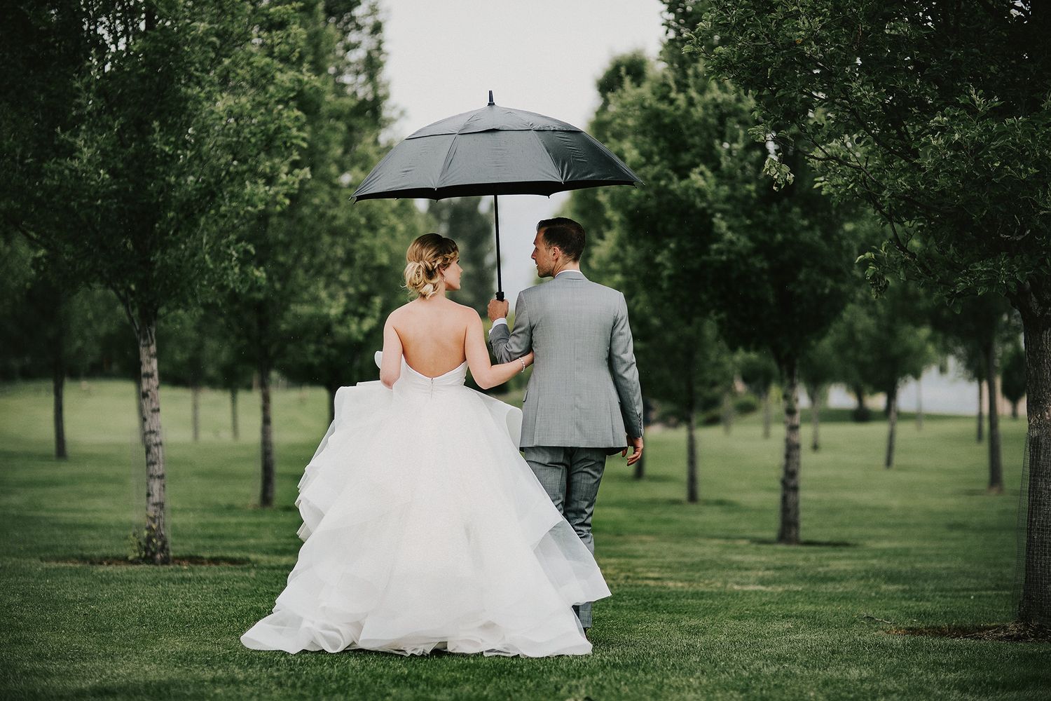 A couple in wedding attire walks together under a black umbrella across a green lawn lined with trees.
