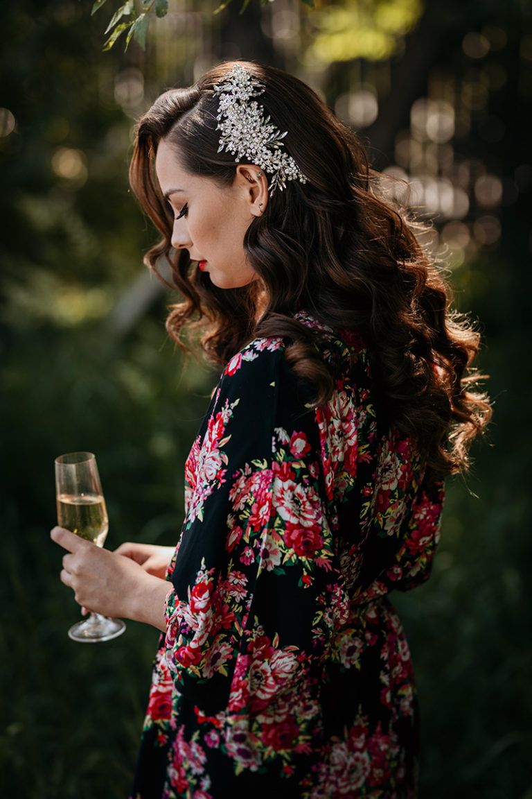 A bride in a black and pink floral bridal robe wearing crystal tiara holds wine glass in outdoor garden setting.