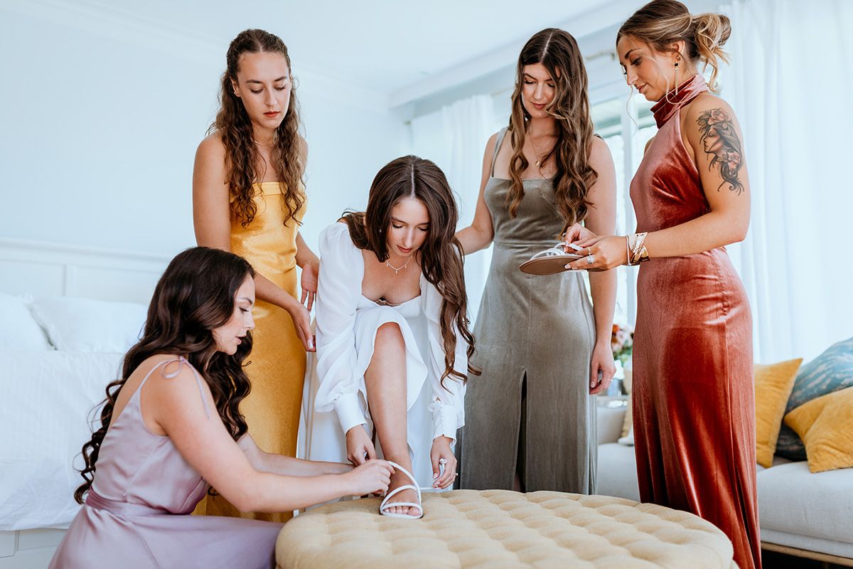 A bridal party in elegant dresses gather around a bride as she adjusts the straps on her shoes.