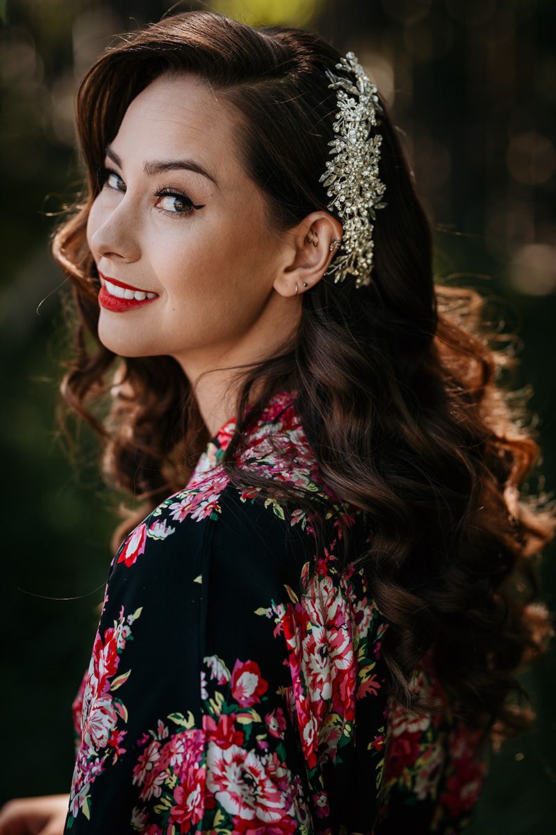 Woman in black floral dress with red roses poses elegantly with delicate white flowers in her dark wavy hair.