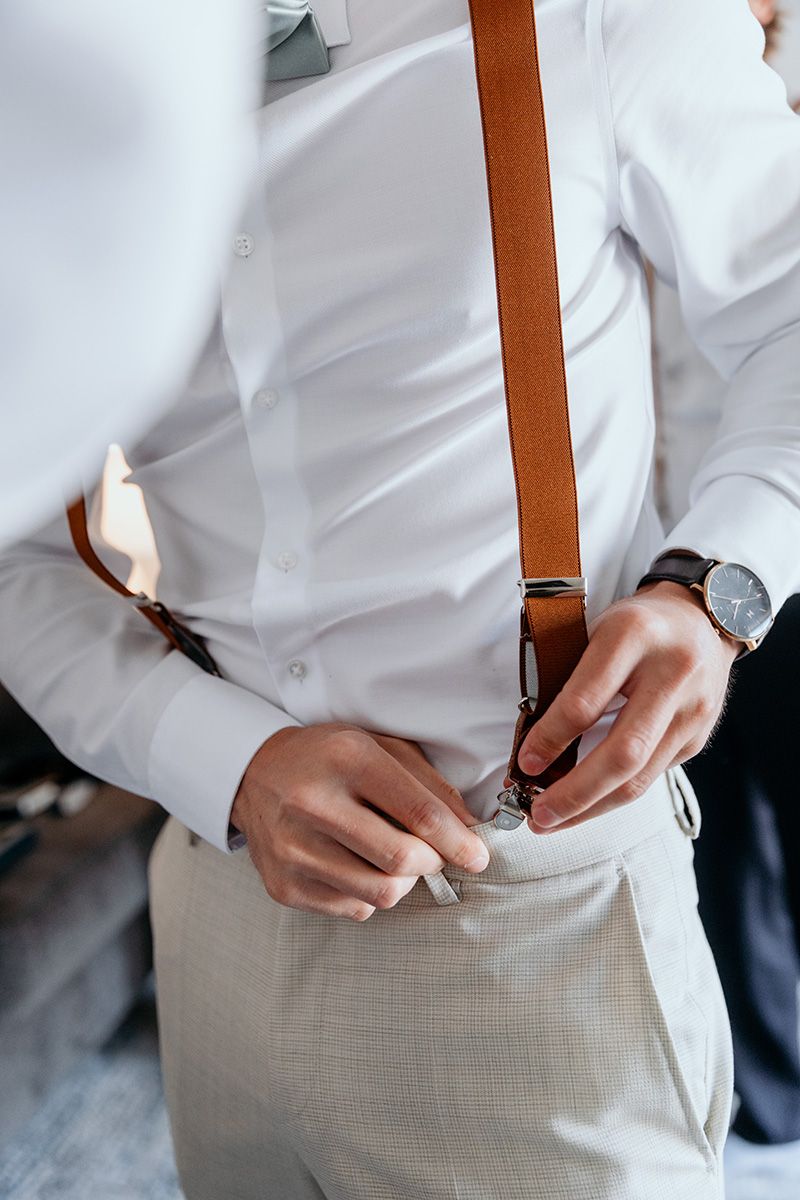 Close-up view of someone wearing a white dress shirt and brown leather suspenders while getting dressed.