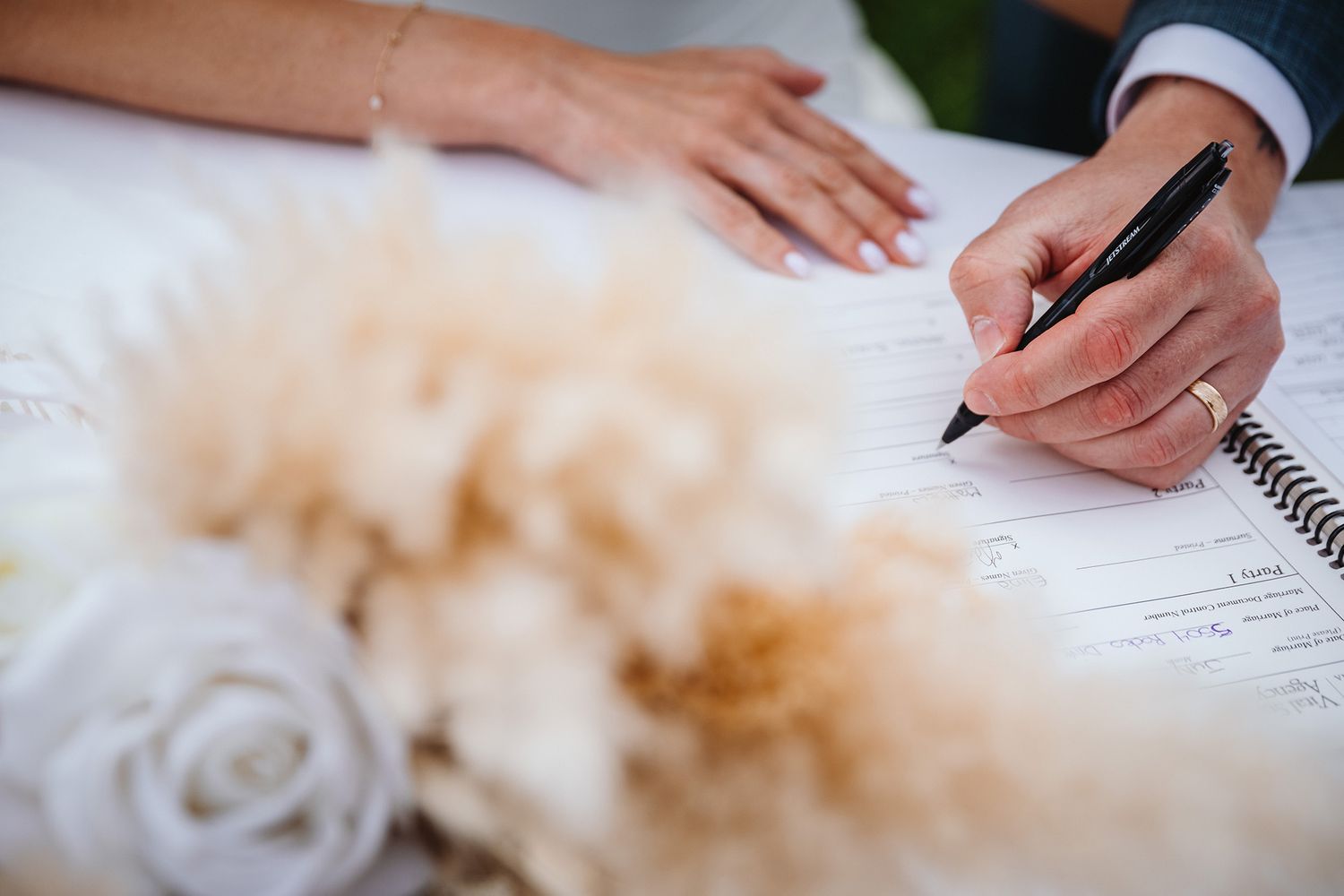 Close-up view of a groom signing in a wedding registry with a pen against a white background.