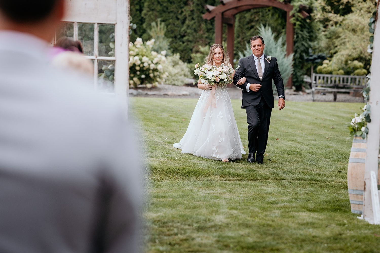 A wedding ceremony takes place on a lush green lawn with guests watching as the bride and her father walk the aisle toward the groom.