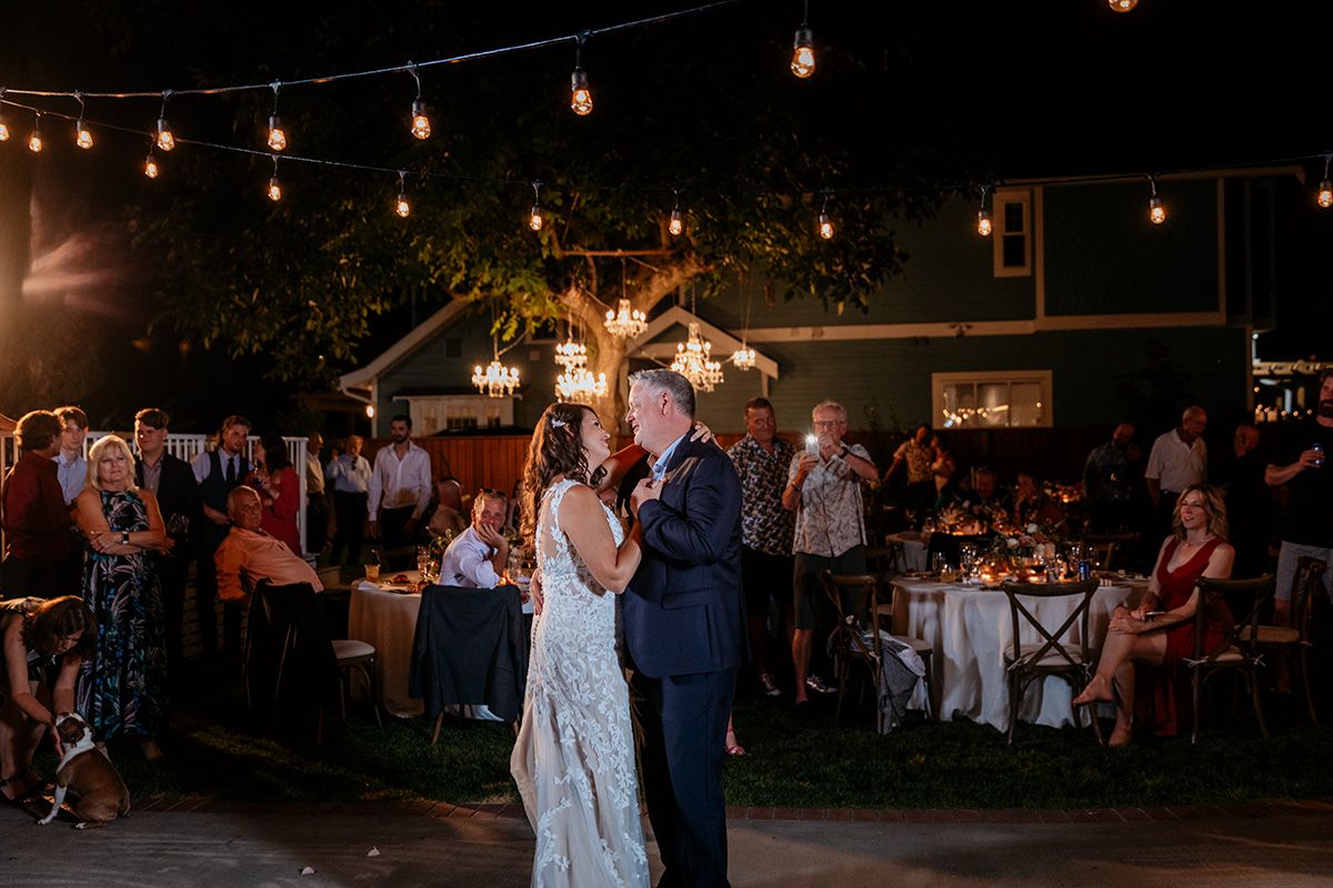 A couple shares their first dance at an outdoor evening wedding reception under string lights and palm trees.