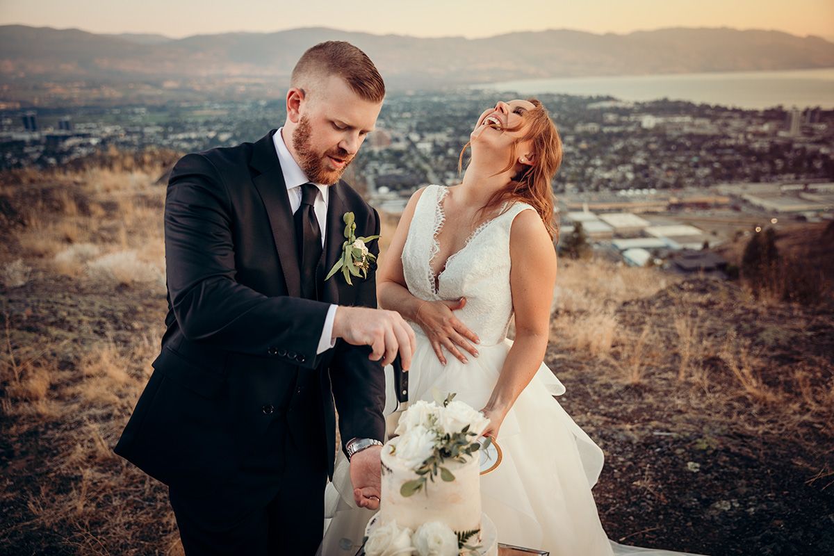 Couple in wedding attire cut their wedding cake at sunset on a mountain overlook with scenic valley views.