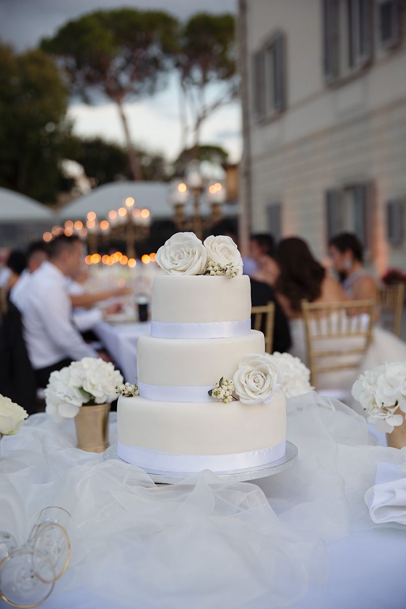 Elegant white wedding cake decorated with white roses sits on a table at an outdoor reception with bokeh lights in background.