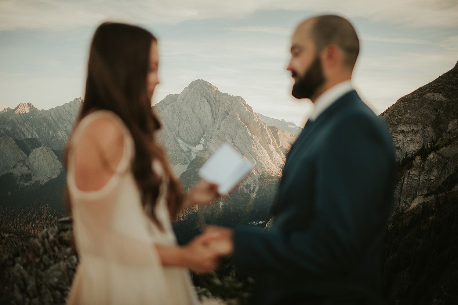 Out of focus in the foreground, a bride shares her vows with her groom against an in focus background of the Canadian Rockies.