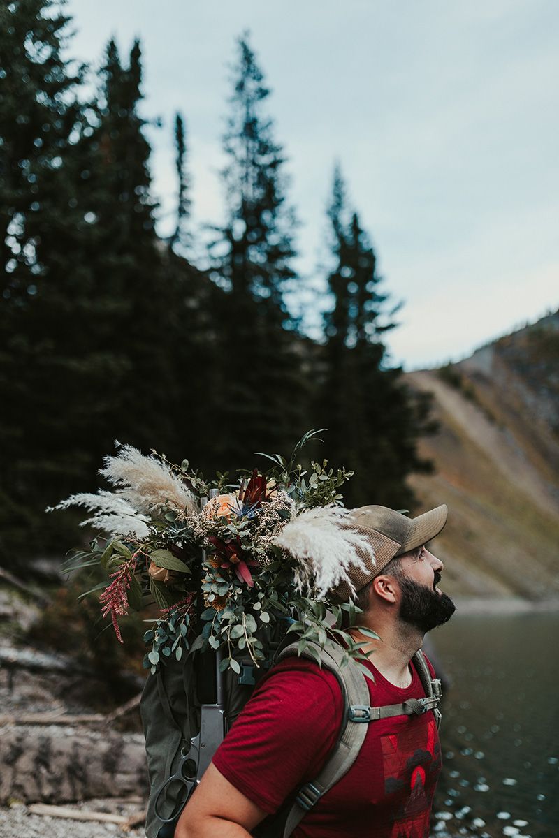 A person wearing a red shirt stands near a mountain lake with evergreen trees and carries a floral backpack arrangement.
