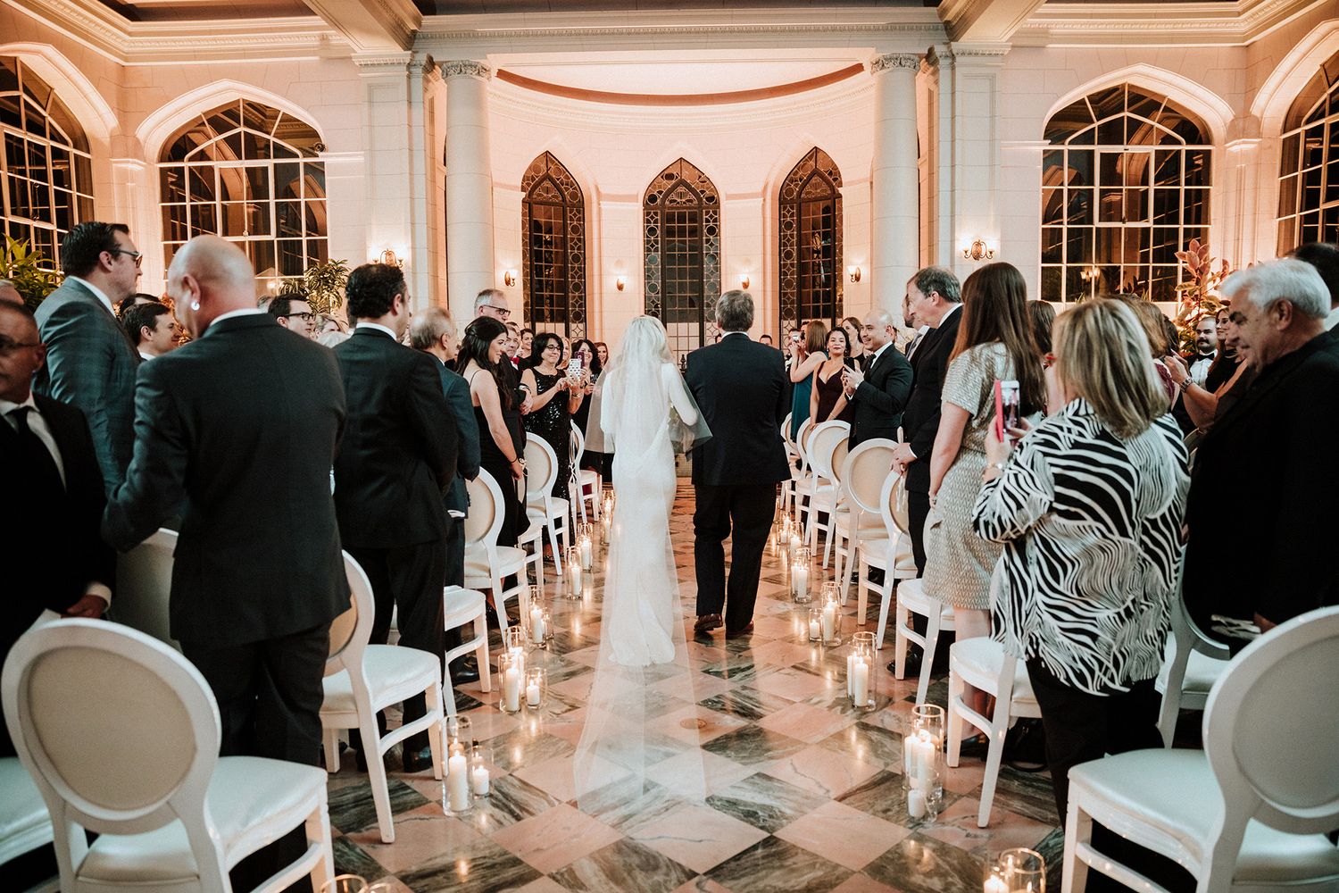 A couple walks down the aisle at an elegant indoor wedding ceremony with white chairs and arched windows.