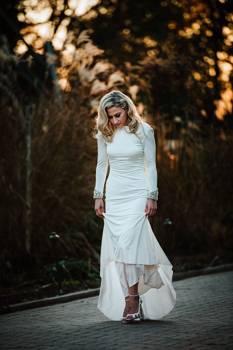 A bride in a flowing white wedding dress poses at sunset on a paved pathway during a golden hour photoshoot.