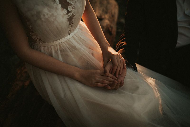 A bride in a flowing white dress stands in soft window light creating an elegant silhouette.