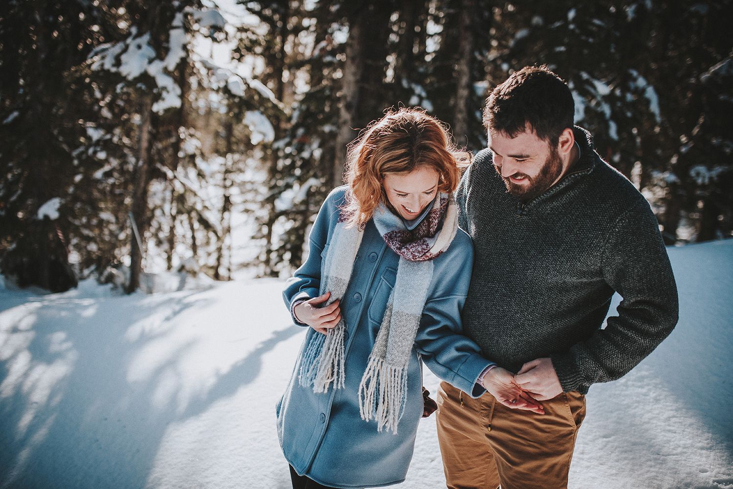 A couple shares a romantic moment in a snowy winter forest setting while holding hands.