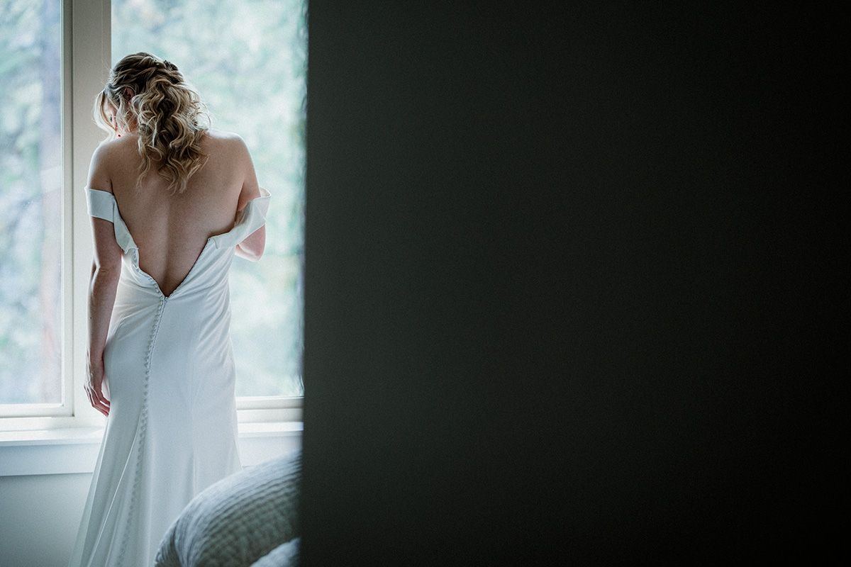 Back view of a bride in an elegant white wedding dress standing by a window in soft natural light.