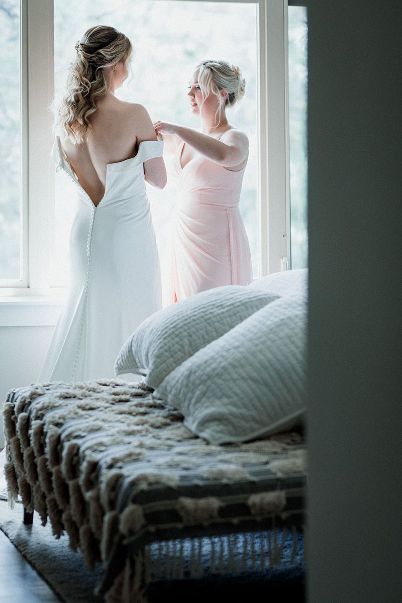 In front of a bright window, a mother helps her bride daughter into her wedding gown.