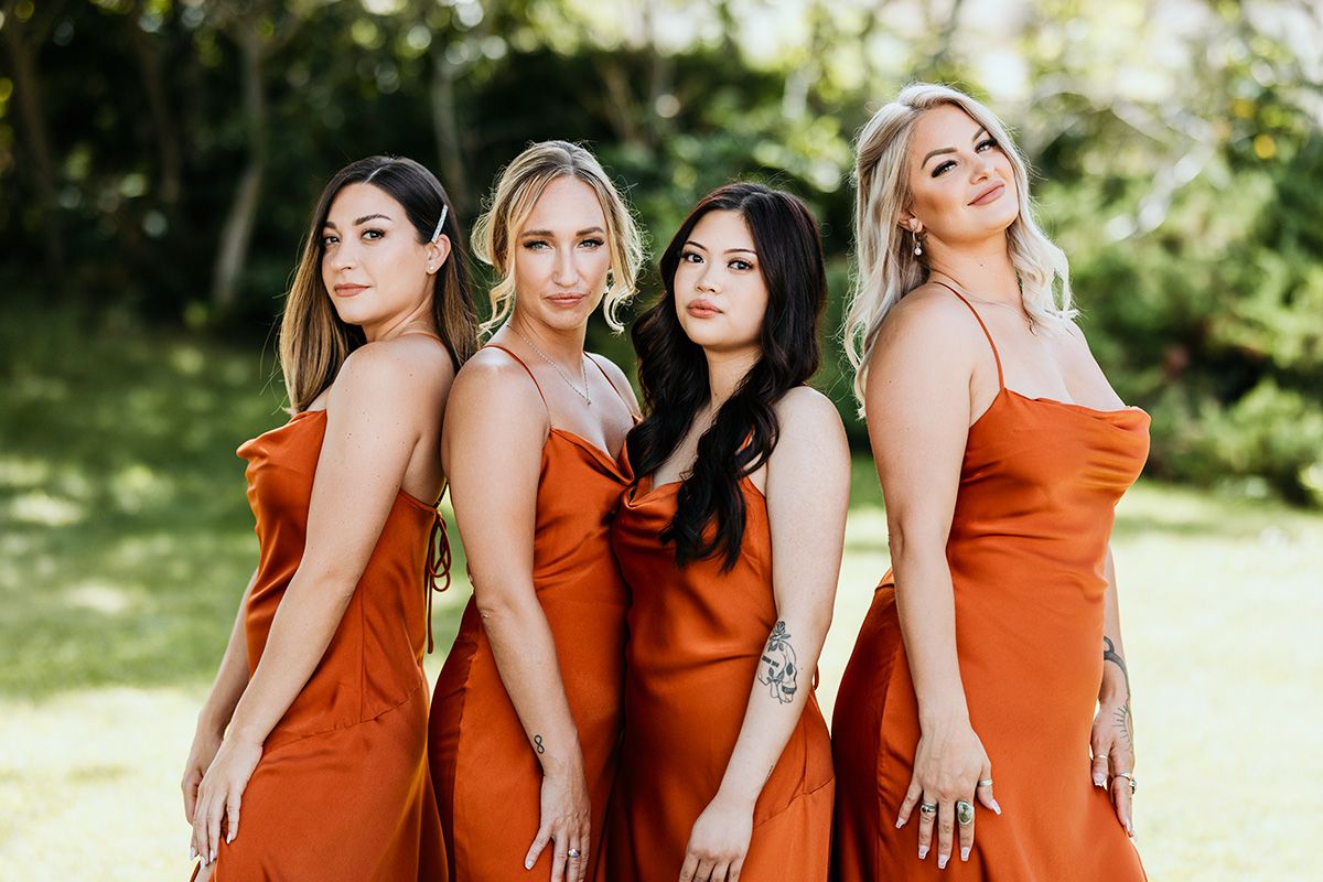 A group of bridesmaids in rust-colored dresses pose together outdoors in natural lighting.
