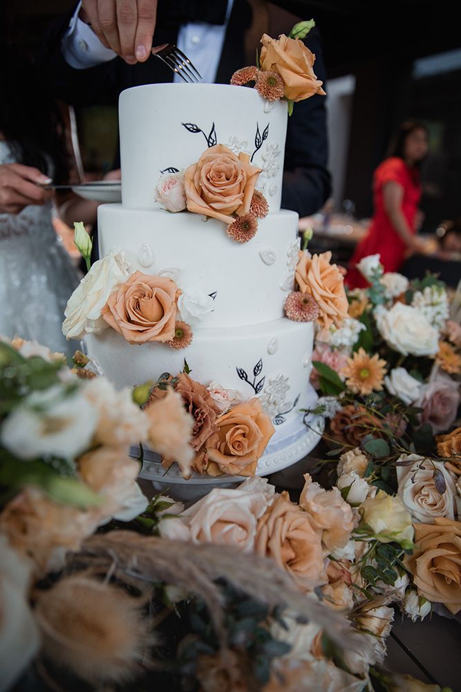 Close-up of elegant white wedding cake decorated with peach and cream roses against dark background.