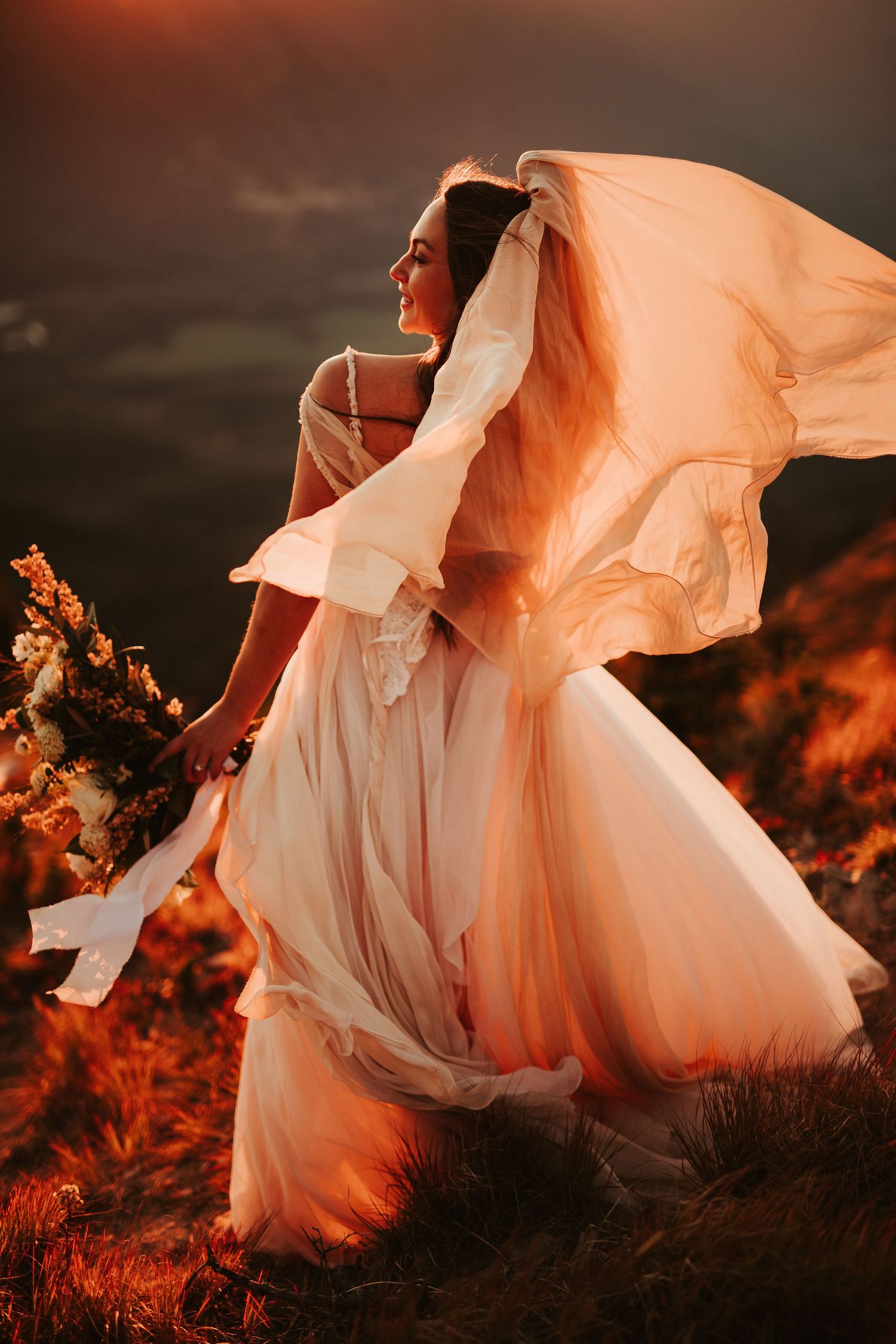 A flowing white wedding dress billows dramatically against a moody sunset landscape creating an ethereal silhouette.