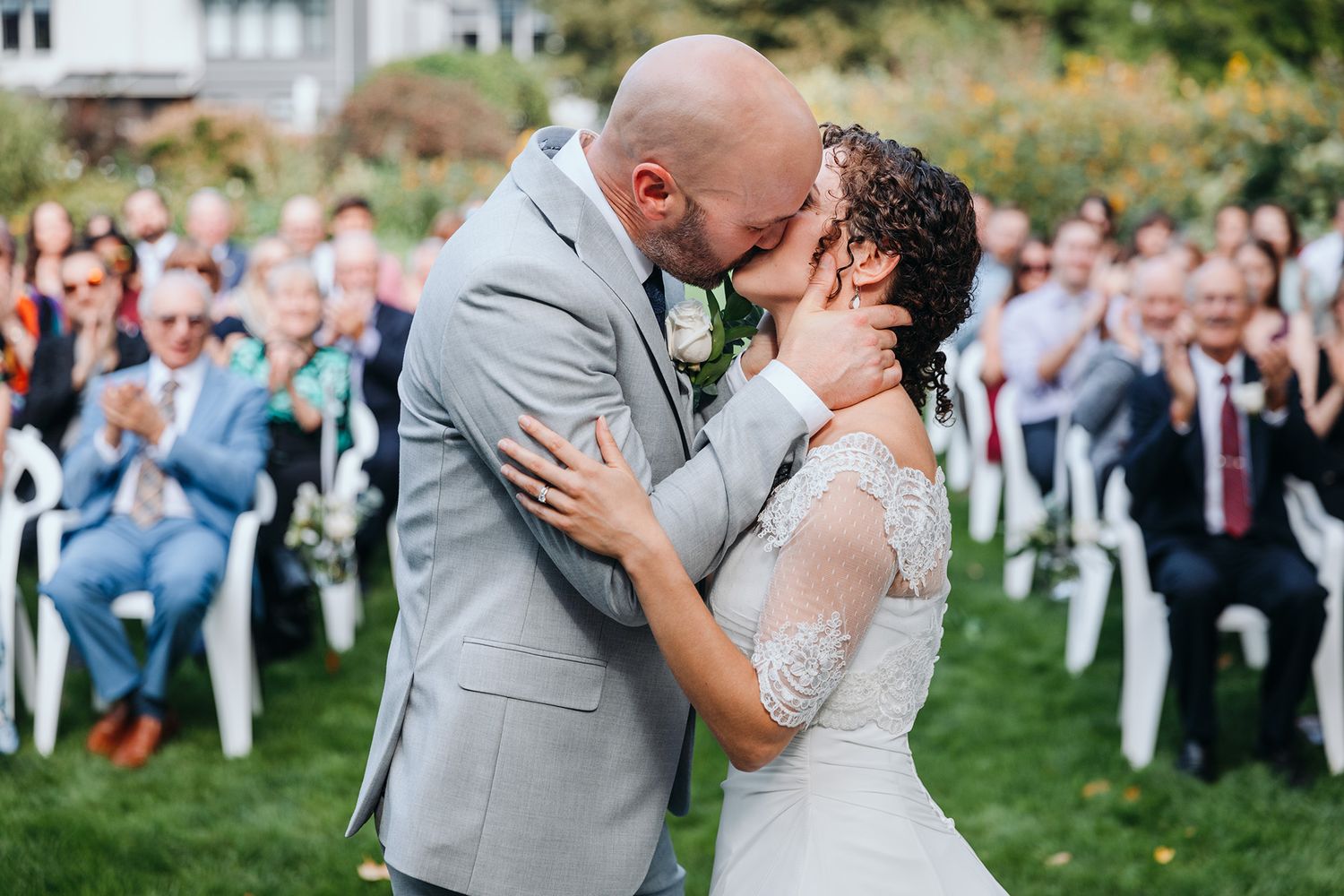 A romantic wedding kiss captured with guests watching in an outdoor garden setting at Guisichan House in Kelowna.
