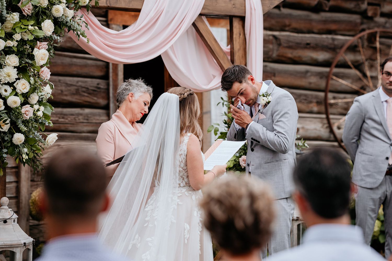 Elegant rustic barn wedding ceremony with pink draping fabric and white floral arrangements. A groom tears up as his bride reads her vows.
