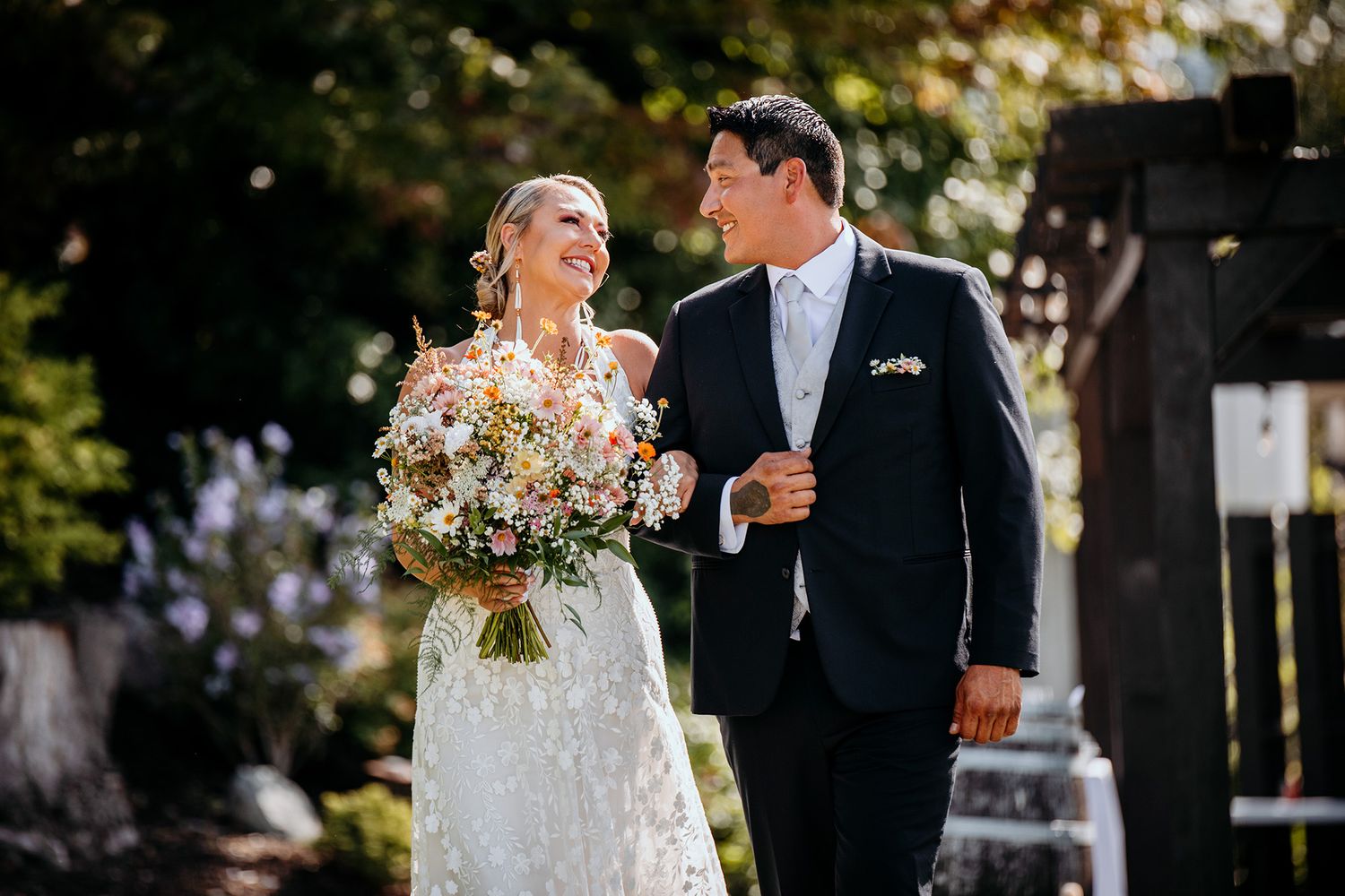 A couple in wedding attire stands together outdoors surrounded by lush greenery and natural lighting.