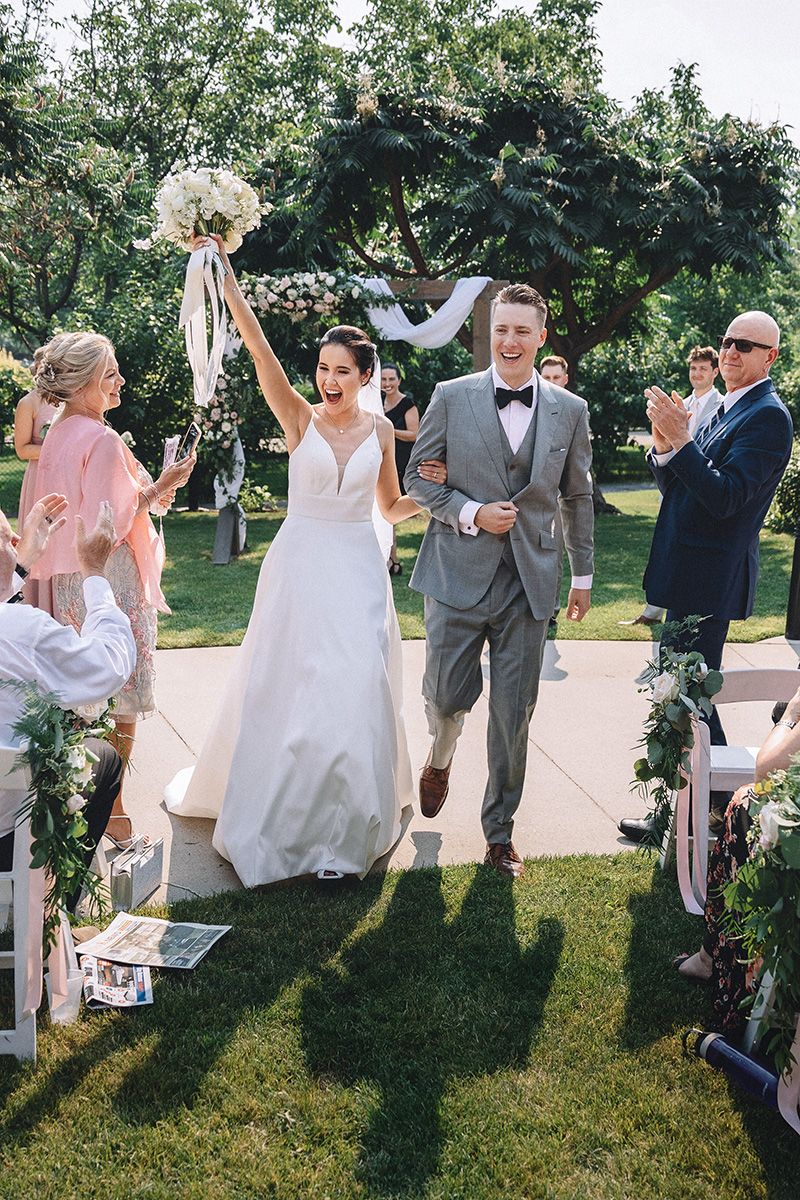 Wedding party celebrates joyfully on a sunny lawn with bridesmaids in blush dresses and groomsmen in grey suits.