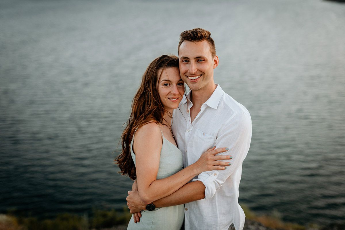 Smiling couple embracing outdoors near water, woman in light mint green dress with long brown hair, man in white shirt.