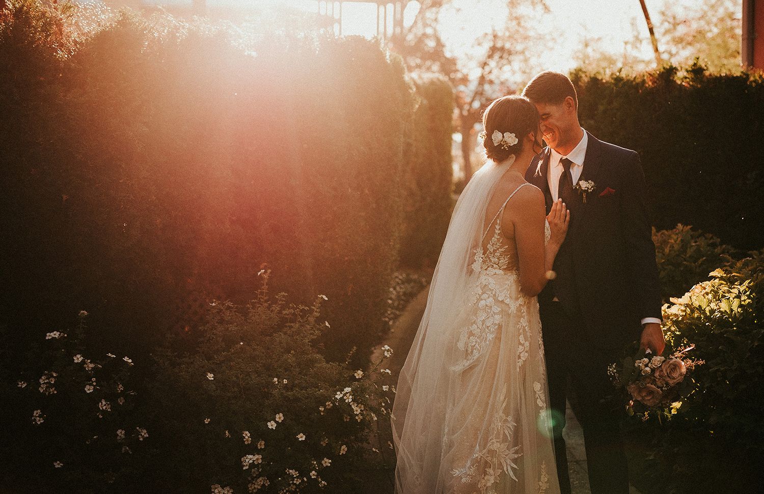 A couple in wedding attire embraces during golden hour in a garden setting with dramatic sunlight filtering through trees.