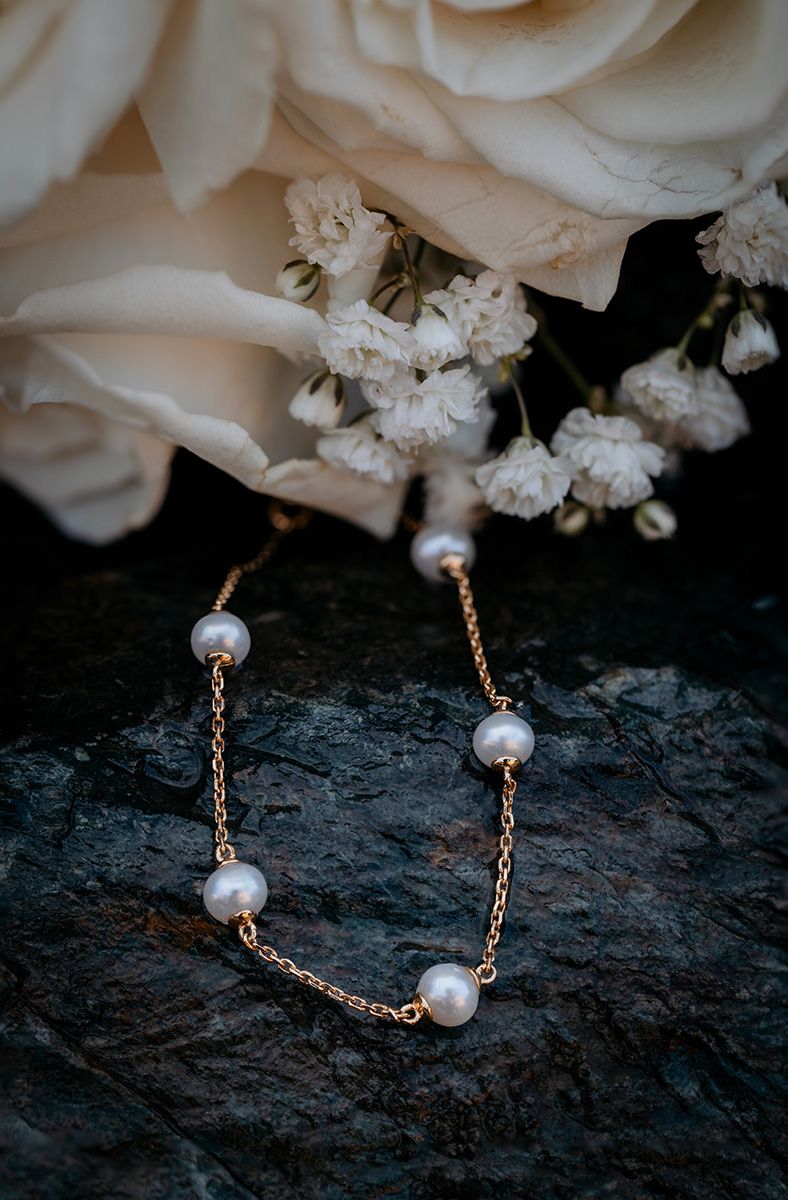 Delicate pearl and rose gold chain bracelet displayed on dark stone with white roses and baby's breath in soft focus background.