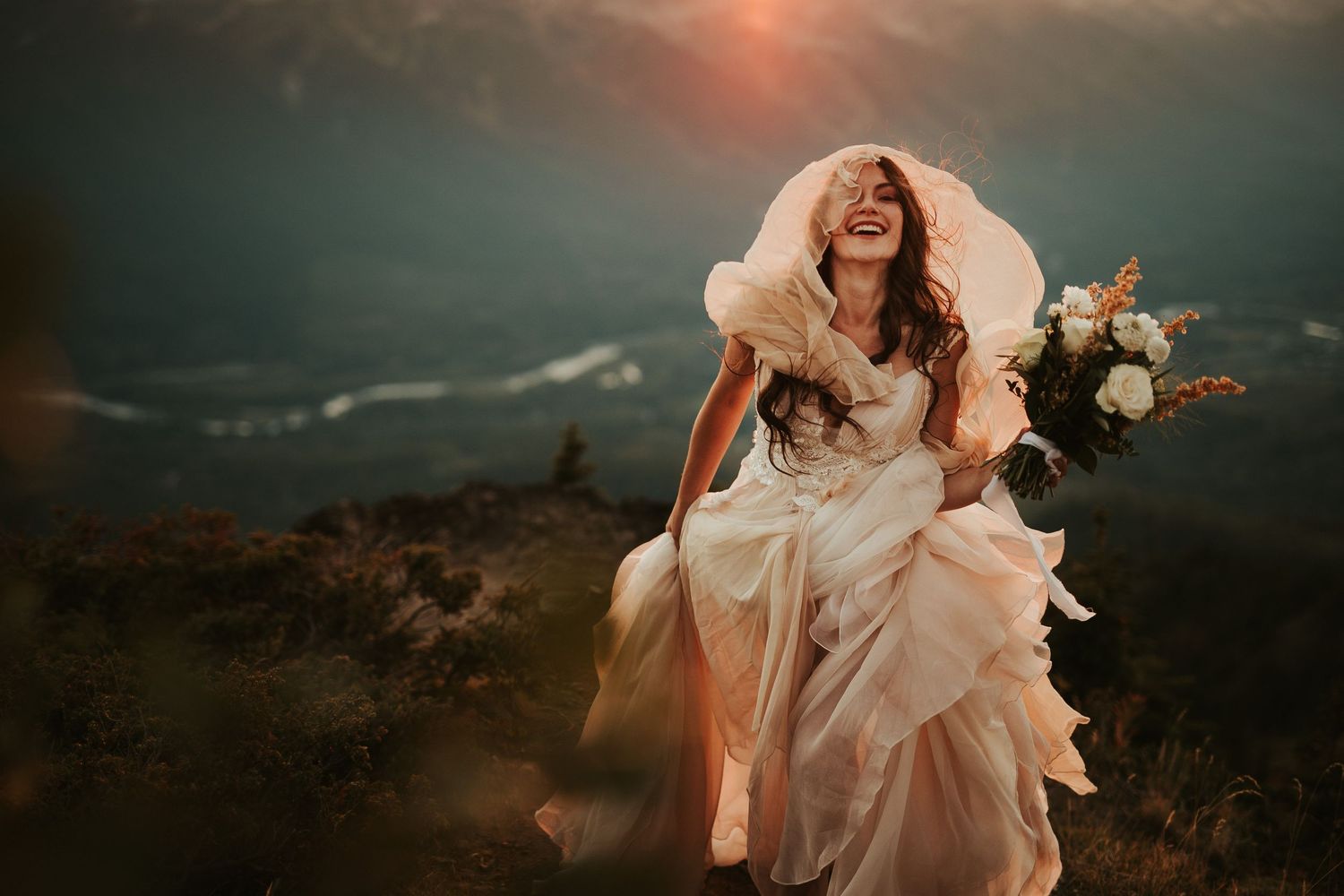 A bride in a flowing white dress runs through a dramatic mountain landscape at sunset. Her dress billows around her head.