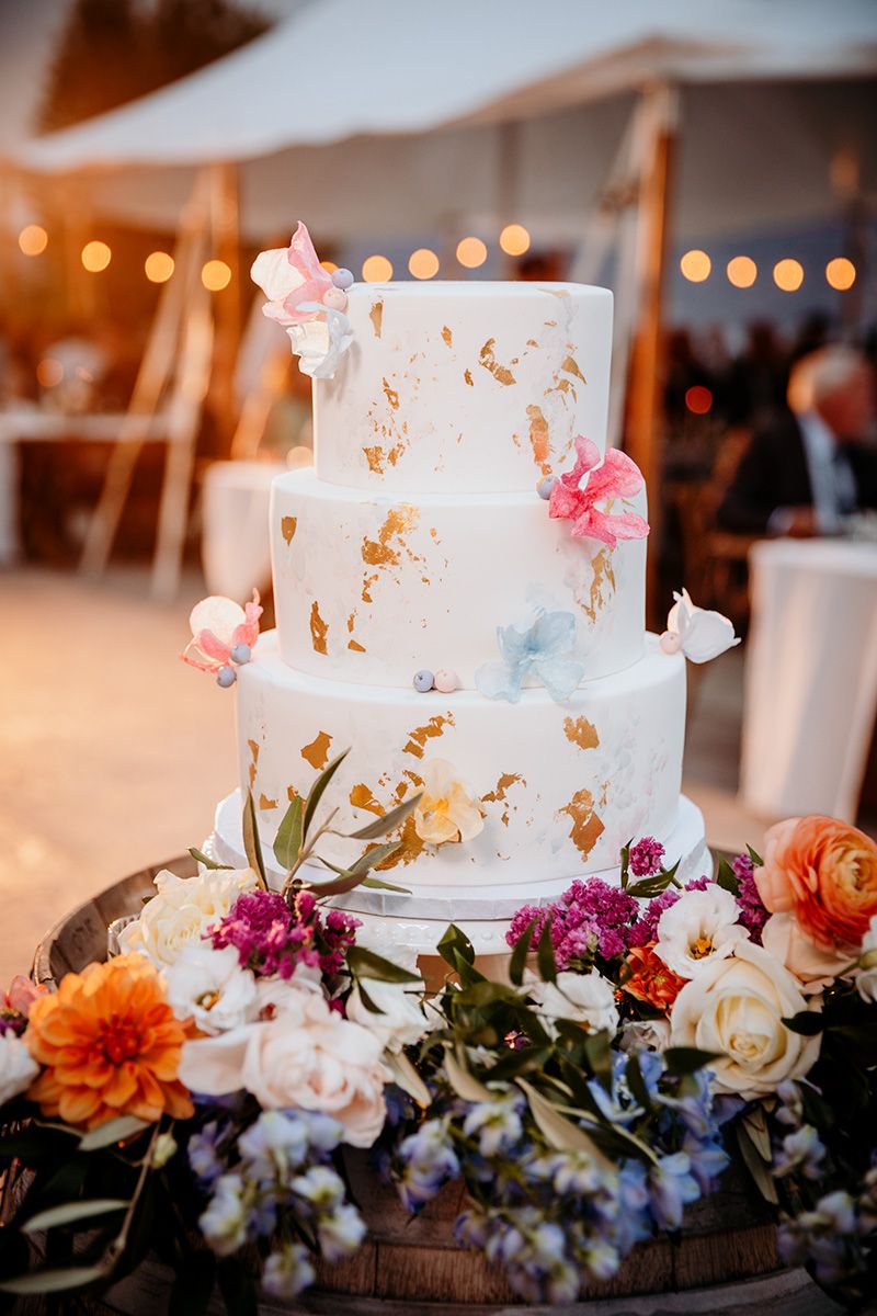White wedding cake adorned with gold leaf and fresh flowers including orchids, roses and dahlias on rustic wood base.