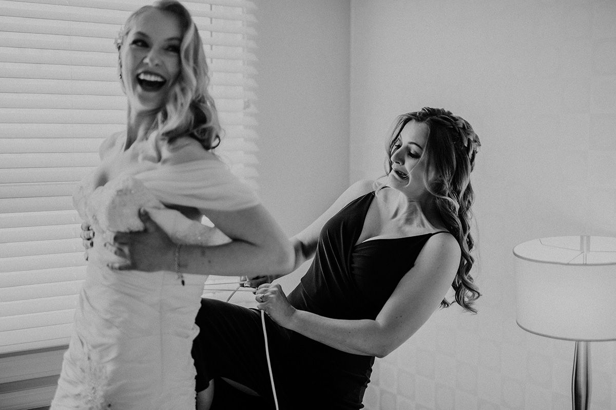 A bride laughs joyfully as her bride's maid puts her knee in the bride's back as she pulls tight the ties of the wedding dress.
