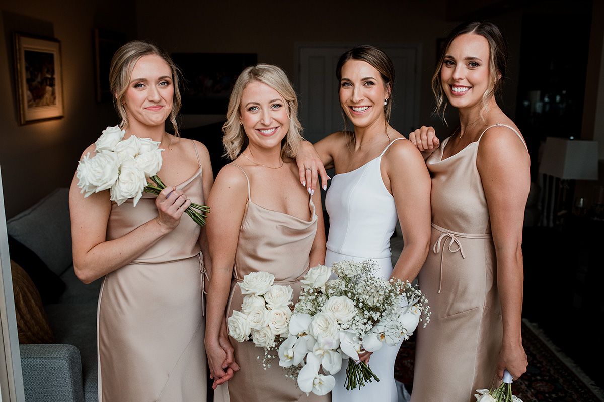 A bridal party wearing champagne-colored satin dresses poses together holding white floral bouquets.