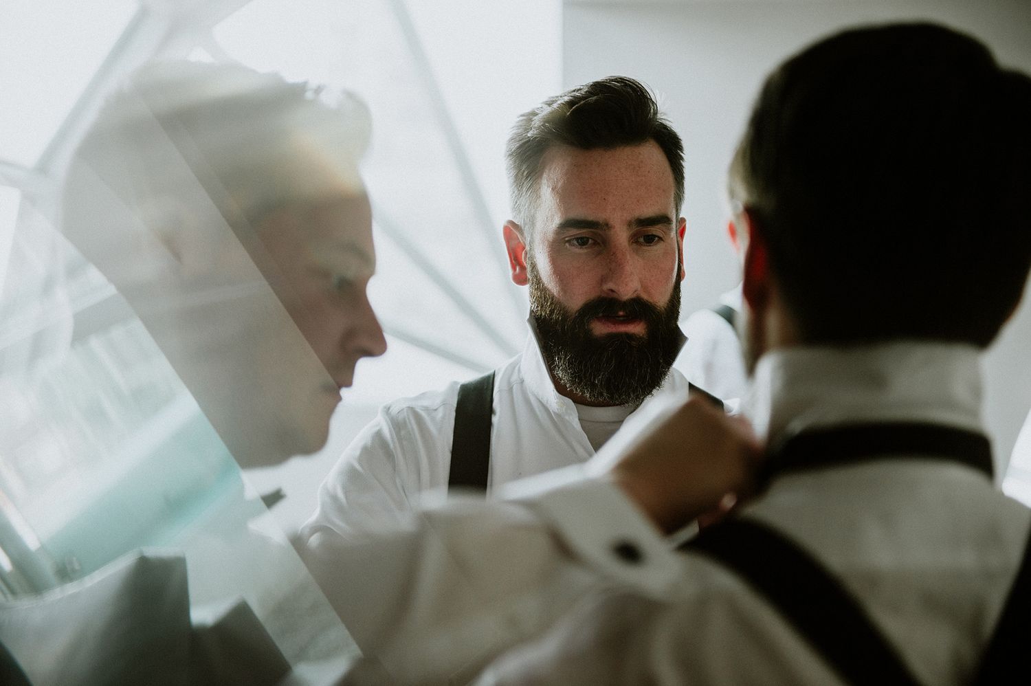 Groomsmen in a bright white room help each other with their ties.