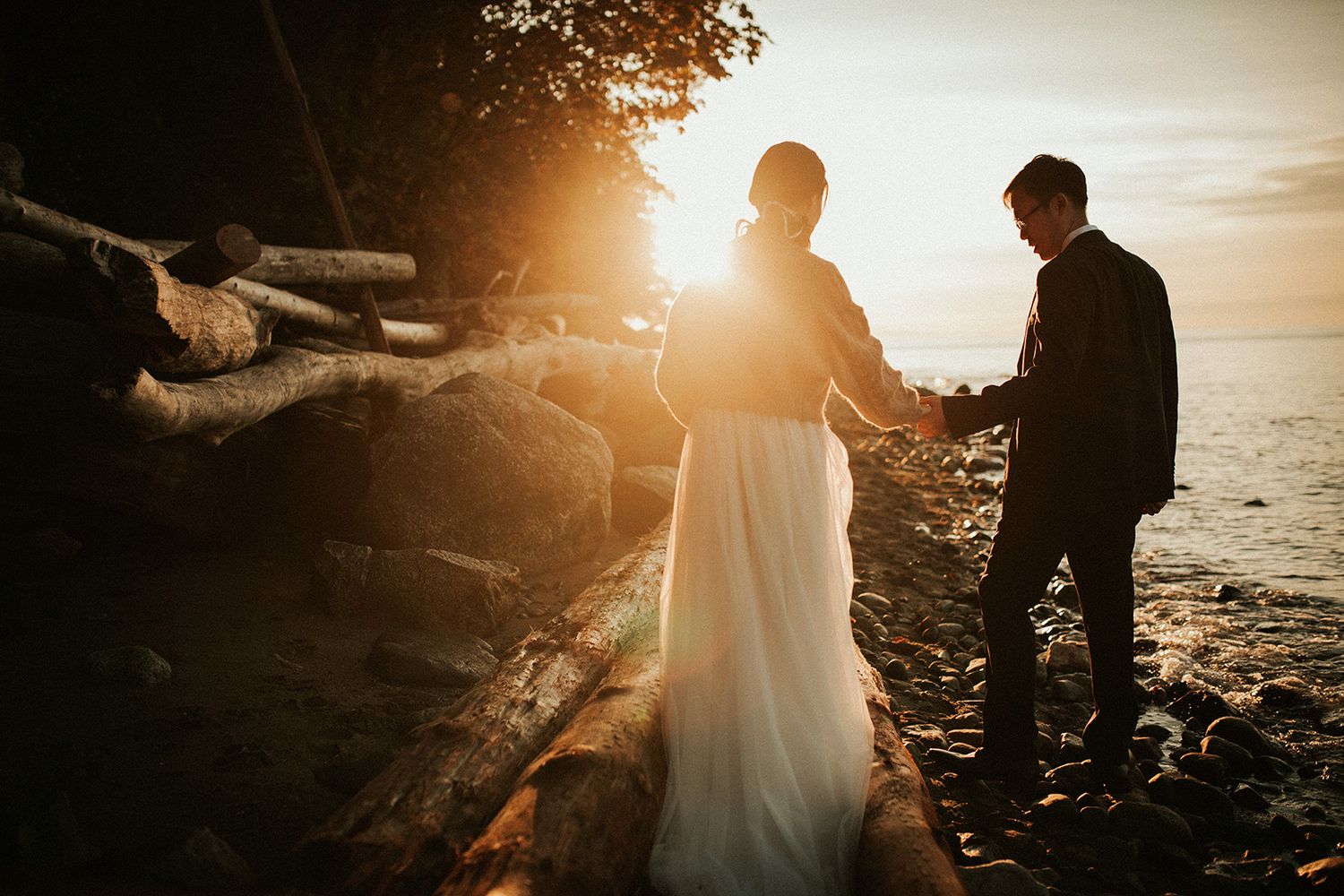 A romantic sunset photo shoot on a rocky beach with golden light illuminating the silhouettes of a groom holding his bride's hand.