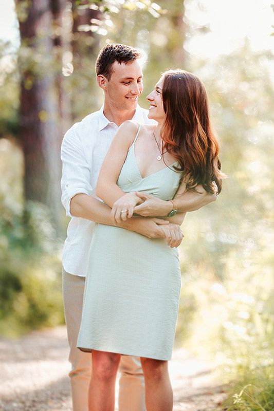 A couple embraces and gazes at each other lovingly on a sunlit forest path, woman in mint dress, man in white shirt.