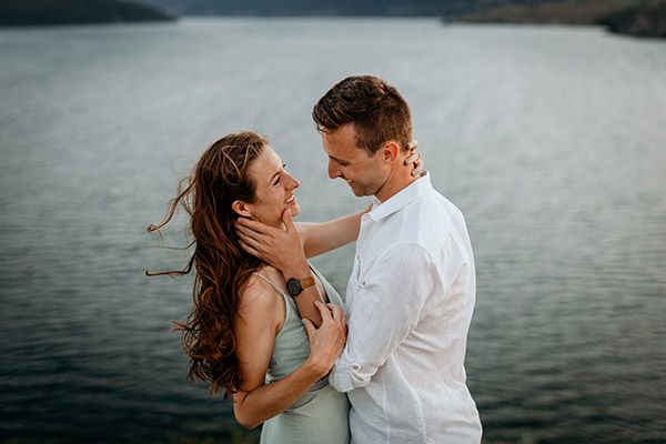 Romantic couple embracing by a serene lake, woman smiling as man holds her face tenderly outdoors.