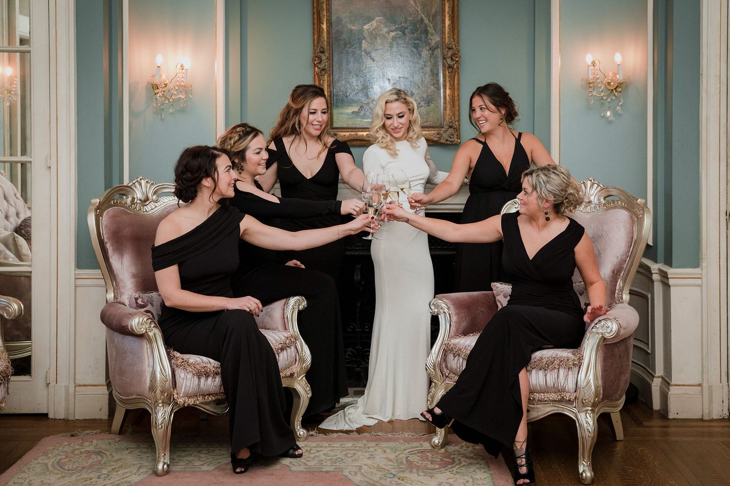 A bridal party in formal attire sitting on elegant vintage furniture share champagne in a light blue room with wall sconces.