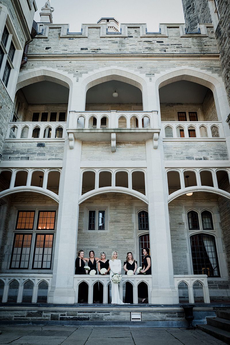 A bridal party poses at the base of an elegant white stone colonnade with arched windows and architectural details at a historic building.