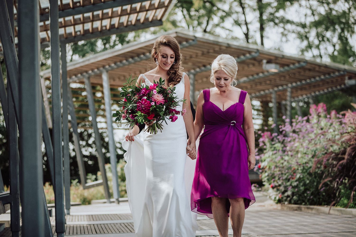 A mother in a purple dress carrying a colorful bouquet walks her bride daughter down the aisle during a wedding ceremony.
