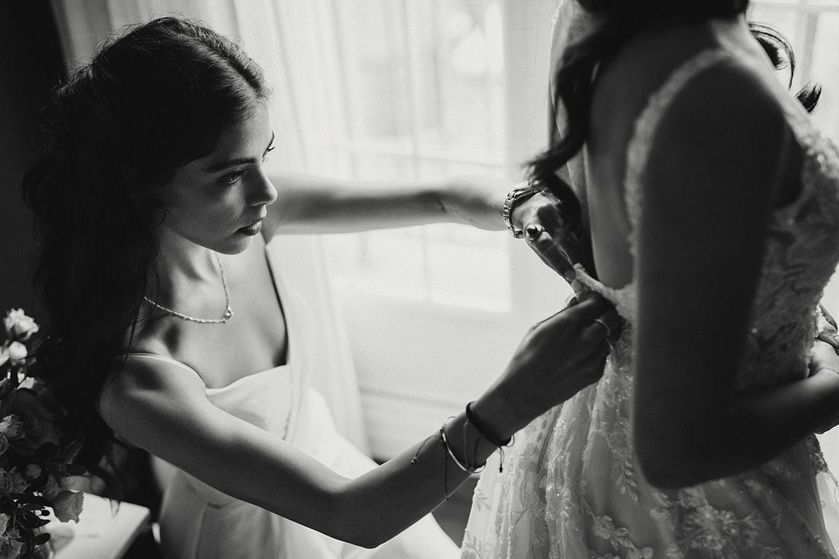 Black and white photo of a woman helping a bride with buttons on the back of her dress.